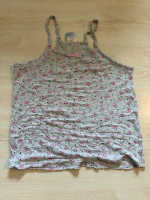 Två linnetoppar med mönster - Säljer två linnetoppar med smala axelband. Den ena är ljusgrå med rosa rosett och små rosa fjärilar, den andra är svart med blommigt mönster i lila och grönt. Perfekta att matcha med jeans eller shorts.