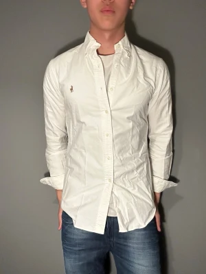 Vit skjorta från Polo Ralph Lauren - Säljer en klassisk vit skjorta från Polo Ralph Lauren med broderad logga på bröstet. Skjortan har långärmad design, knappar framtill och är gjord i mjuk bomull. Perfekt för dig som vill ha en stilren och tidlös look.
