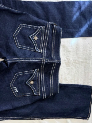 Mörkblå lågmidjadejeans från Guess - Snygga mörkblå jeans från Guess med bootcut passform och vita sömmar.  Stängbara fickor baktill med Sten-detaljer. Midja tvärs över: 37 cm Innerbenslängd: 83 cm