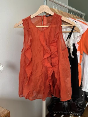 Orange volangtop från Zara - Säljer en snygg orange ärmlös topp från Zara med volangdetaljer framtill. Toppen har en lätt transparent look och är tillverkad i ett tunt, luftigt material som passar perfekt till varma dagar. 