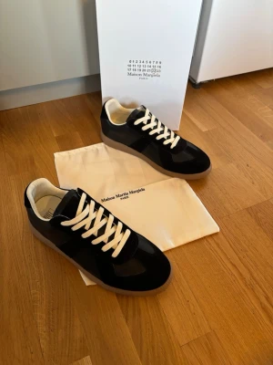 Maison Margiela svarta sneakers - Maison Margiela sneakers i svart läder och mocka med vita skosnören och beige innersida. Klassisk low-top modell med gummisula och stilren design. Perfekt för dig som vill ha en ikonisk och trendig sneaker.