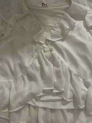 Vit volangblus med omlott och knappar - Säljer en romantisk vit blus med volanger och omlott-design. Blusen har knappar framtill och är tillverkad i ett lätt, transparent material som ger ett luftigt intryck. Perfekt för dig som gillar feminina detaljer och en elegant look.