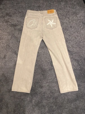 Corteiz jeans - Säljer ett par gråa jeans från corteiz med raka ben och unika broderade detaljer på bakfickorna – en stjärna och en corteiz märket. Klassisk femficksmodell med läderpatch i midjan. Perfekta för dig som vill sticka ut med coola detaljer och streetwear-vibe.