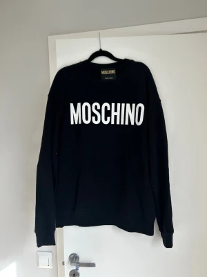 Svart hoodie från Moschino - Snygg svart hoodie från Moschino med stor vit logga framtill. Klassisk passform med rund hals och långa ärmar. Perfekt för dig som gillar streetwear och vill ha något stilrent men ändå statement.