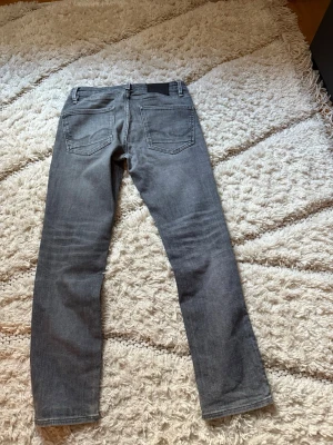 Grå jeans från Jack n Jones - Säljer ett par grå jeans från Nudie Jeans med raka ben och klassisk femficksmodell. Jeansen har tydliga sömmar på bakfickorna och en svart patch vid linningen. Materialet är denim i bomull med en mjuk tvättad look.