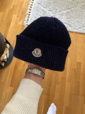 Mörkblå ribbstickad Moncler mössa - Säljer en mörkblå ribbstickad mössa från Moncler med klassisk uppvikt kant och broderad Moncler-logga framtill. Mössan är tillverkad i mjukt material och har en tidlös design som passar perfekt till kalla dagar.