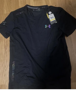 Under Armour svart träningsset - Säljer ett svart träningsset från Under Armour med t-shirt och shorts. T-shirten har ett färgglatt logotryck på bröstet och diskret text längs sidan. Shortsen har elastisk midja med snörning och logotyp på benet. Perfekt för gym eller löpning.