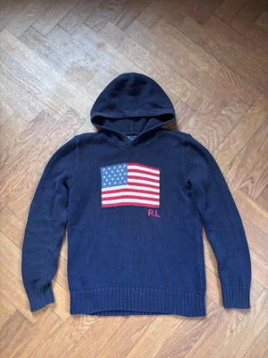Marinblå stickad hoodie Polo Ralph Lauren - Stickad marinblå hoodie från Polo Ralph Lauren med amerikansk flagga i rött, vitt och blått på bröstet samt RL-broderi. Tröjan har ribbstickade muddar och är tillverkad i mjukt material. Perfekt för dig som vill ha en cool och avslappnad look. USA