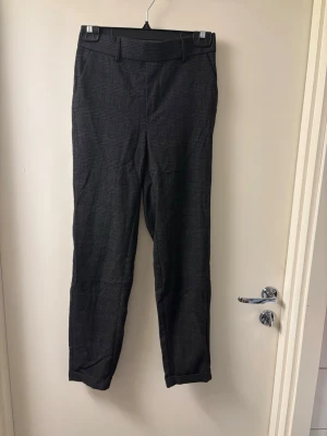Mörkgrå kostymbyxor med rutig design - Snygga mörkgrå kostymbyxor med diskret rutig mönster och klassisk passform. Byxorna har hällor för bälte, fickor fram och elastisk midja bak för extra komfort. Perfekta för en stilren och modern look.