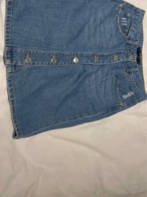 Blå jeanskjol med knappar framtill - Snygg blå jeanskjol i kort modell med knappar längs hela framsidan. Kjolen har klassiska fickor och en lätt A-linjeformad siluett. Perfekt till vardags för en avslappnad och trendig look.