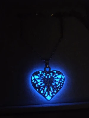 Halsband med lysande hjärthänge - Unikt handgjort halsband med silverfärgad kedja i rostfritt stål och hjärtformat hänge. Hänget har ett fjärilsmotiv och lila detaljer som lyser blått i mörker efter att vanligt dagsljus eller solljus har nått hänget en stund. Bär halsbandet som vanligt så sköter sig effekten självt. Lyser vanligtvis i flera timmar & kan lysa upp till 12 timmar. Perfekt för dig som gillar smycken med lite extra wow-faktor och snygga detaljer.