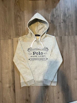 Grå hoodie från Polo Ralph Lauren - Snygg grå hoodie från Polo Ralph Lauren med stor mörkblå logga och text på bröstet🤗 använd ett par gånger men inga fel på den🙌🏻 inget äkthetsbevis tyvärr. Hör av er vid snabbaffär samt funderingar🤩