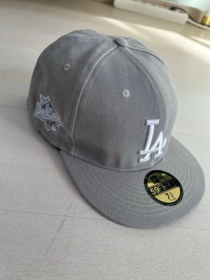 Grå LA New Era keps - Snygg grå keps från New Era 59Fifty med LA Dodgers-logga broderad i vitt framtill och detaljer på sidan. Klassisk flat brim och strukturerad passform. Tillverkad i bomullstyg och perfekt för dig som gillar streetwear och sportig stil. Storlek 7 3/4 eller 61.5cm