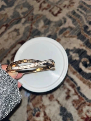 Guldigt armband med romerska siffror - Snyggt guldigt armband med tvinnad design där ena delen är dekorerad med små glittrande stenar och den andra har ingraverade romerska siffror. Perfekt accessoar för att lyfta din outfit med lite extra bling.