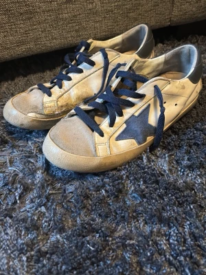 Golden Goose skor - Säljer ett par klassiska Golden Goose sneakers i vitt skinn med mörkblå snörning och en ikonisk blå stjärna på sidan. Hälpartiet är också blått och skon har en vintage-inspirerad look. Perfekta för dig som gillar streetstyle och unika detaljer. lite slitna på insidan men går enkelt att limma