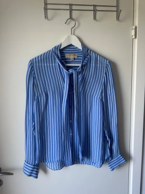 Blåvit randig blus med knytband Michael Kors - Snygg blå blus med vita vertikala ränder från Michael Kors. Blusen har långa ärmar, knappar framtill och ett dekorativt knytband vid halsen. Tillverkad i ett mjukt, lätt material som ger en avslappnad men ändå chic look. Storlek S. Använt tre gånger 🩷