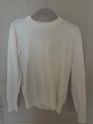 Vit stickad tröja med rund hals - En enkel och stilren vit stickad tröja med rund hals och långa ärmar. Tröjan har ribbstickade muddar vid hals, ärmslut och nederkant. Perfekt att matcha med jeans eller kjol för en clean look. Har fina detaljer vid axlarna som en liten scrunch som är väldigt fin nu för varmare tider med en kjol men även för höst med ett par jeans