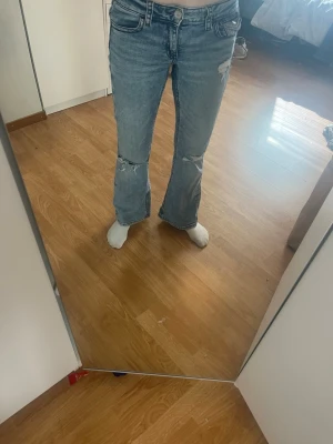Blå bootcut jeans med slitningar - Säljer ett par ljusblå bootcut jeans med slitningar på knäna och en liten trasig detalj vid fickan. Jeansen har klassiska fem fickor och är gjorda i denim. 