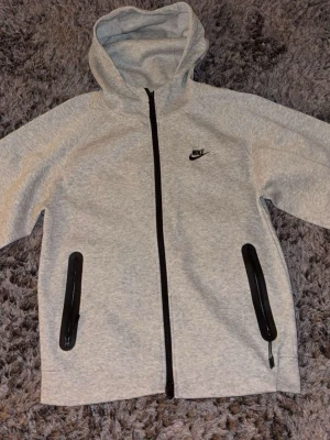 Nike tech kofta  - Snygg ljusgrå hoodie från Nike med hel dragkedja framtill och svarta detaljer. Hoodien har två fickor med dragkedja och en klassisk huva. Nike-logga broderad på bröstet. Perfekt för en sportig och avslappnad stil.