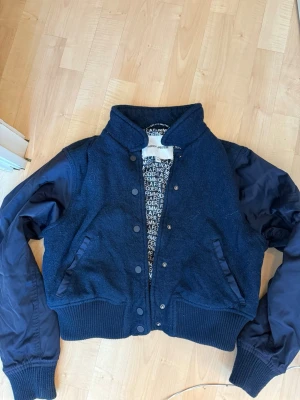 Mörkblå overshirt från Carin Wester/ålens  - Snygg mörkblå overshirt från Carin Weater med ullblandad kropp och glansiga ärmar i nylon. Jackan har ribbade muddar, tryckknappar framtill och två fickor. Fodret har ett coolt textmönster. Perfekt för lager-på-lager och streetstyle.