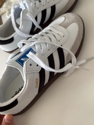 Adidas Samba vita och svarta sneakers - Säljer ett par klassiska Adidas Samba sneakers med vit läderöverdel, svarta ränder och grå mockadetaljer. Skorna har bruna gummisulor och blå Adidas-logga på tungan. Perfekt för dig som gillar retrostil och ikoniska modeller.