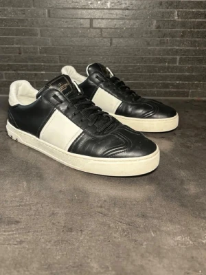 Svarta och vita sneakers från Valentino - Snygga sneakers från Valentino med svart och vitt läder, klassisk låg modell och svarta skosnören. Tungan har en svart logotypdetalj och sulan är vit med subtila nitar på hälen. Perfekt för dig som vill ha en stilren och exklusiv look.