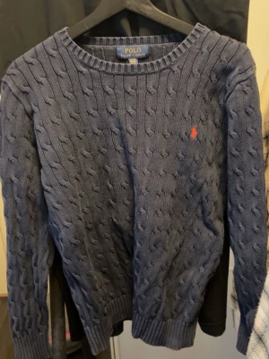 Polo Ralph Lauren / Cotton Cable Pullover Tröja - Kabelstickad blå tröja från Polo Ralph Lauren med rund halsringning och den klassiska röda logotypen broderad på bröstet. Tröjan har ribbade muddar vid ärmslut och nederkant, perfekt för en stilren och avslappnad look. Storlek 170/88A (Small)