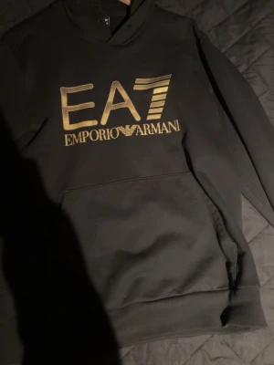 Svart hoodie från Emporio Armani EA7 - Snygg svart hoodie från Emporio Armani EA7 med stort guldfärgat logotryck på bröstet. Klassisk passform med huva och känguruficka framtill. Perfekt för dig som gillar exklusiva streetwear-vibes och vill sticka ut med ett lyxigt märke.