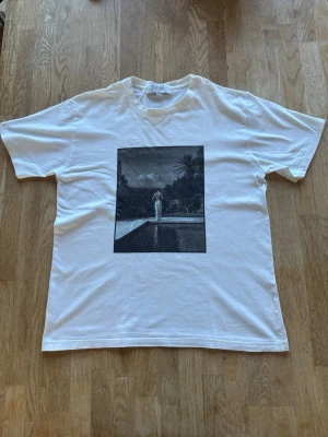 Vit t-shirt med svartvit print från Fiore studios - Vit t-shirt från Fiore studios med en stor svartvit print på framsidan som föreställer en person vid en pool omgiven av palmer. T-shirten har klassisk passform och rund hals. Perfekt för dig som gillar stilrena motiv och enkel design.