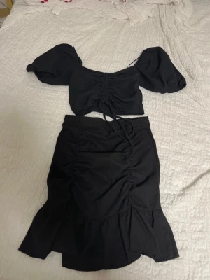 Svart omlott set från Gina Tricot - Säljer ett svart set från Gina Tricot med puffärmad croppad topp och matchande kjol. Toppen har omlottdetalj och snörning framtill, kjolen har volangkant och rynkade detaljer. Perfekt för dig som gillar trendiga och feminina plagg.