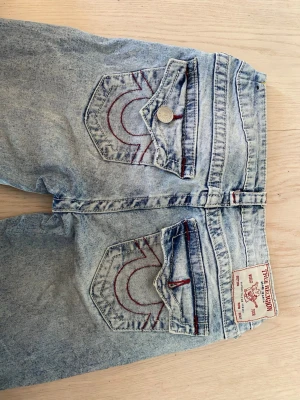 True Religion Jeans - Säljer ett par ljusa blå bootcut jeans från True Religion w24 med klassiska röda sömmar och snygga bakfickor, jeansen är uppsydda så passar någon som är ungefär 155 - 160 cm, ena knappen har lossnat på ena bakfickan därav priset (se bild 1 & 3), men inget man tänker på när dom är på , skitsnygga!! 💕💕Nypris: 1200kr