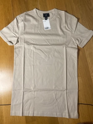 Beige slim fit t-shirt från H&M - Snygg beige t-shirt från H&M i slim fit-modell. Tillverkad i mjuk bomull med klassisk rund hals och korta ärmar. Perfekt basplagg för en clean och enkel stil.