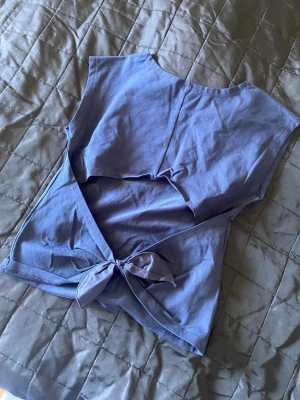 Blå omlott topp med knyt - Säljer en blå omlott topp i mjuk bomull. Toppen är ärmlös och har ett snyggt knyt framtill som ger en cool och avslappnad look. Perfekt att matcha med jeans eller kjol för en trendig stil.