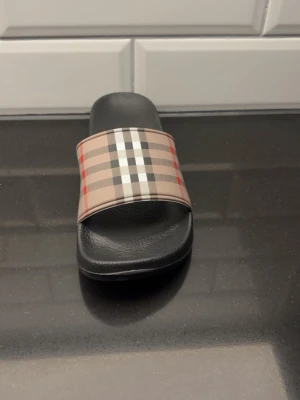 Burberry rutiga slides i svart - Säljer ett par snygga slides från Burberry med klassiskt beige, svart, vitt och rött rutmönster över foten. Sulan är svart och har en bekväm formad design i syntetmaterial. Perfekta för en trendig och avslappnad stil.