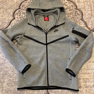 Grå hoodie från Nike med dragkedja - Grå hoodie från Nike med svarta detaljer och hel dragkedja framtill. Tröjan har huva, lång ärm och en praktisk ficka med dragkedja på ena ärmen. Materialet är mjukt och känns sportigt, perfekt för chill eller träning. Nike-logga på bröstet och ärmen. Har tyvärr inte prislappen kvar kan skicka bättre bilder.
