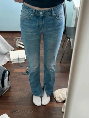 Blå raka jeans med slits framtill - Säljer ett par ljusblå jeans med raka ben och slits framtill vid bensluten. Jeansen har klassisk femficksmodell, hög midja och knappgylf. Perfekta för en avslappnad och trendig look. 