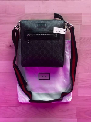 Svart axelväska från Gucci - Snygg svart axelväska från Gucci med klassiskt GG-mönster, justerbar axelrem i svart och rött samt en praktisk ytterficka med dragkedja. Väskan är i ett tåligt material och har en stilren, fyrkantig form. Perfekt för dig som vill ha en trendig och lyxig accessoar.