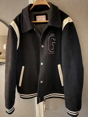 Svart bomberjacka från Guess - Snygg svart bomberjacka från Guess med vita detaljer på axlar och fickor. Jackan har broderad G-logga och texten 'Los Angeles' på bröstet. Ribbstickade muddar och krage ger en klassisk varsity-look. Perfekt för dig som vill ha en stilren och trendig jacka.