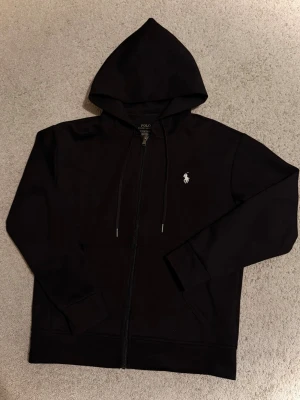 Polo Ralph Lauren tröja - Säljer en svart hoodie från Polo Ralph Lauren med dragkedja och huva. Klassisk logga broderad i vitt på bröstet. Hoodien har ribbade muddar och fickor framtill. Perfekt för dig som gillar stilren design och premiumkänsla.