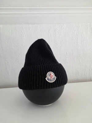 Svart ribbstickad mössa - Svart ribbstickad mössa med uppvikt kant och klassiska märkes logga framtill. Mössan har en enkel, stilren design och är tillverkad i ett mjukt, värmande material som passar perfekt för kyliga dagar.