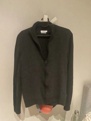 Mörkgrå ziptröja från mario conti - Mörkgrå ziptröja från Mario conti med hög krage och hel dragkedja framtill. Tröjan har lång ärm och är tillverkad i ett mjukt stickat material som ger en clean och stilren look. Perfekt att slänga på sig över en t-shirt.