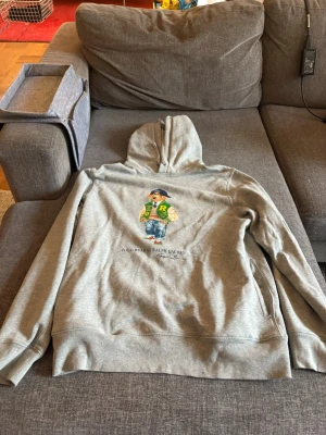 Grå hoodie Polo Bear Ralph Lauren - Säljer en grå hoodie från Ralph Lauren med den ikoniska Polo Bear-trycket på ryggen. Björnen har en grön collegejacka, jeans och sneakers. Hoodien har huva och är gjord i mjukt bomullsmaterial. Perfekt för dig som gillar streetwear och klassisk stil.