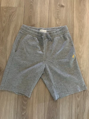 Grå mjukisshorts från Pier One - Grå mjukisshorts från Pier One med elastisk midja och snörning. Shortsen har två sidofickor fram och en bakficka. Liten gul logga broderad på vänster ben. Perfekta för chill eller träning.
