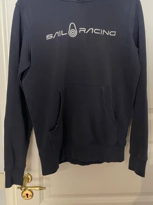 Marinblå Sail Racing hoodie - Snygg marinblå hoodie från Sail Racing med vit logga på bröstet. Klassisk känguruficka framtill och huva baktill. Tillverkad i mjukt bomullsmaterial som känns skönt mot huden. Perfekt för dig som gillar stilren och sportig look.