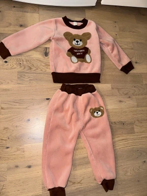 Rosa loungewear-set med nallemotiv - Mysigt tvådelat loungewear-set i mjuk rosa fleece med bruna muddar. Tröjan har ett stort nallemotiv framtill och byxorna har en liten nalle på ena benet. Perfekt för hemmamys och chill. Sött och bekvämt set för dig som gillar gulliga detaljer.