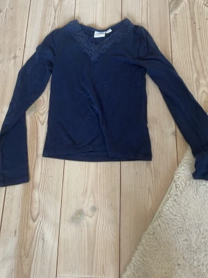 Mörkblå långärmad topp Gina Tricot - Mörkblå långärmad topp från Gina Tricot med v-ringning och spetsdetaljer vid halsen. Tillverkad i mjukt material som känns skönt mot huden. Perfekt för dig som gillar stilrena plagg med en liten twist.