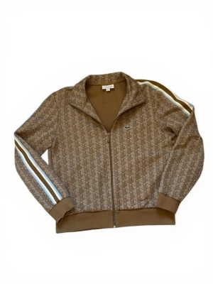 Lacoste monogram - Snygg brun ziptröja från Lacoste med diskret mönster och klassisk krokodil-logga på bröstet. Tröjan har ribbade muddar, ståkrage och vita samt ljusblå ränder längs ärmarna. Regular fit och tillverkad i mjukt material som känns skönt mot huden. Helt ny, använd två gånger.