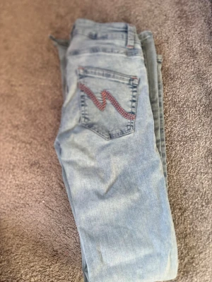 Ljusblå slitna bootcut jeans - Säljer ett par ljusblå bootcut jeans med slitningar och hål på båda knäna. Jeansen har klassisk femficksdesign och en cool detalj med röd zigzag-söm på bakfickan. Tillverkade i denim, perfekta för en avslappnad streetstil.