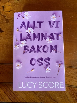 Allt vi lämnat bakom oss - En skönlitterär bok av Lucy Score med lila omslag dekorerat med vita blommor. Boken är tredje delen i succéserien Knockemout och handlar om passion, hemligheter och förflutna som jagar huvudpersonerna Lucian och Sloane.