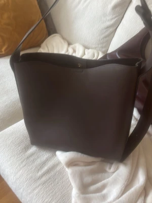 Mörkbrun axelväska i läderlook - Stilren mörkbrun axelväska med bred axelrem och minimalistisk design. Väskan har en slät yta och rymlig insida, perfekt för att bära med sig det viktigaste. Passar dig som gillar enkel och modern stil.
